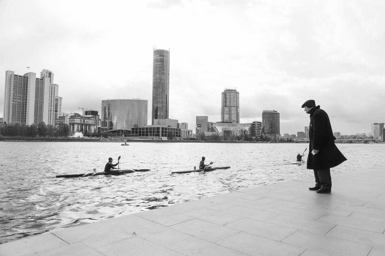 Фотография сделана на городском пруду во время соревнований по гребле на байдарках. Было довольно холодно, несмотря на конец мая. Наблюдая за спортсменами, я обратила внимание на этого дедушку. Он уже замерз, но что-то никак не давало ему уйти. Что-то щемящее было в том, как он смотрит на ребят в байдарках, как провожает их взглядом, словно провожает свою молодость, и как ветер раздувает его пальто …
